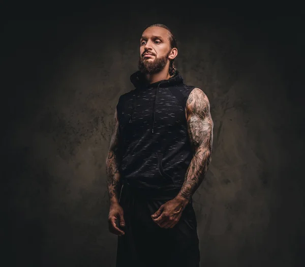 Retrato de un atleta tatuado barbudo con capucha negra. Aislado sobre un fondo de textura oscura . — Foto de Stock