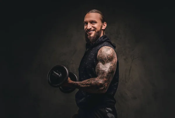 Sorrindo atleta tatuado barbudo em um capuz preto posando com um halteres na mão. Isolado em fundo escuro texturizado . — Fotografia de Stock