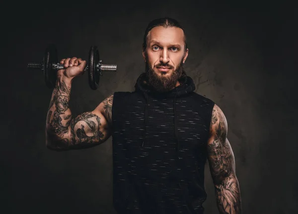 Retrato de um atleta tatuado barbudo em um capuz preto posando com um halteres na mão . — Fotografia de Stock