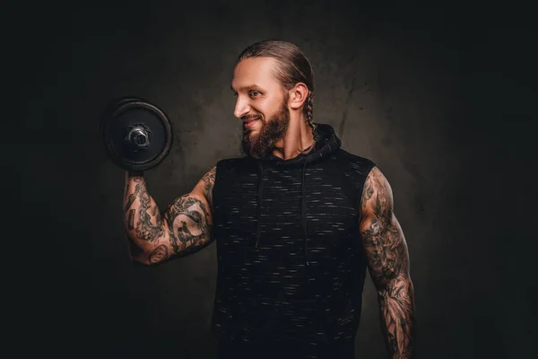 Sorrindo atleta tatuado barbudo em um capuz preto posando com um halteres na mão. Isolado em fundo escuro texturizado . — Fotografia de Stock