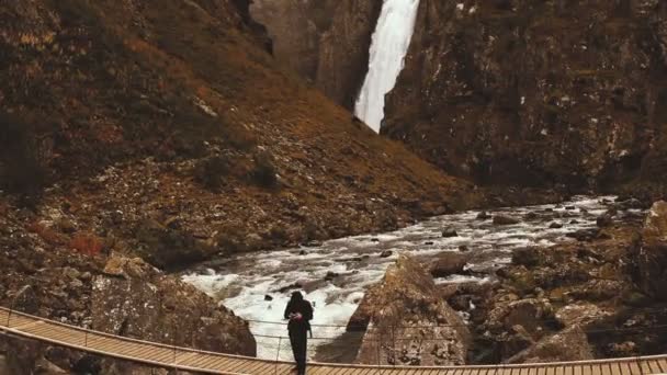 Nature photographer getting shot while standing on narrow wooden bridge against a beautiful waterfall in autumn Norway. — Stock Video