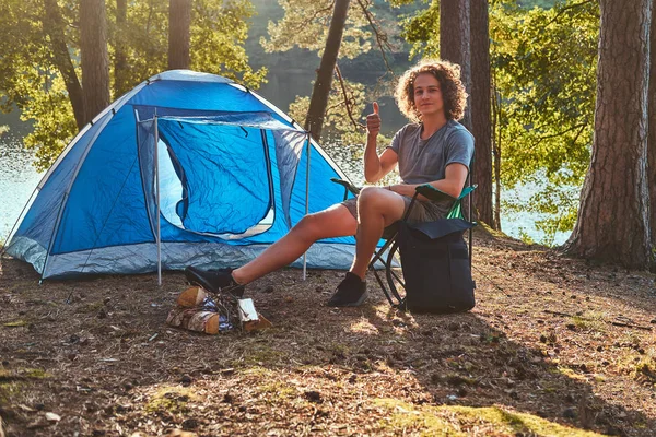 Fiatal természetjáró fickó ül egy széken, és remek felbukkan az erdő Camp göndör haj. — Stock Fotó