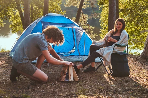 Joven pareja de excursionistas en el campamento en el bosque. Joven intenta encender un fuego . —  Fotos de Stock