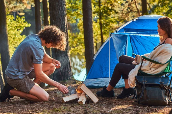 Joven pareja de excursionistas en el campamento en el bosque. Joven intenta encender un fuego . — Foto de Stock