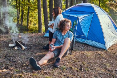 Genç çift oturan yürüyüşçü orman kampında bir kamp ateşi yakın güneşli günde ısınma.