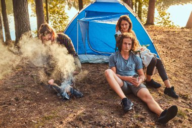 Üç arkadaş ormanda ve kampta bir ateş yakıp çalışın hiking. Seyahat, turizm ve zammı kavramı.