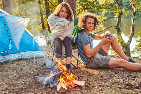 Jovem casal de caminhantes sentados aquecendo perto de uma fogueira no acampamento na floresta no dia ensolarado . — Fotografia de Stock