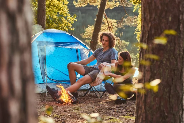 Unga par vandrare sitter uppvärmningen nära en lägereld på camp i skogen på solig dag. — Stockfoto