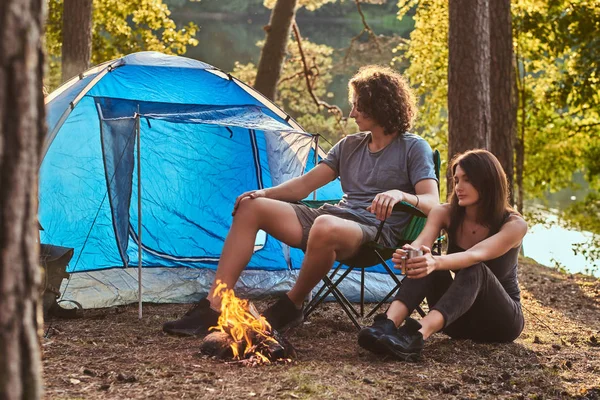 Et ungt par fotturister som sitter ved bålet i skogen på solskinnsdag. . – stockfoto