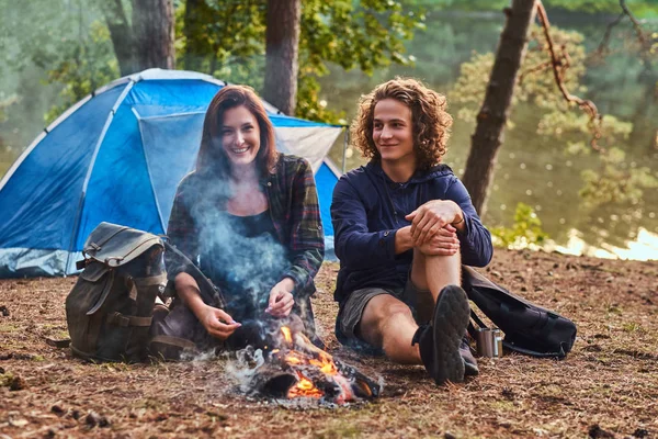 Joven pareja de excursionistas sentados y hablando mientras se calientan cerca de una fogata en el campamento en el bosque. Viajes, turismo y concepto de caminata . — Foto de Stock