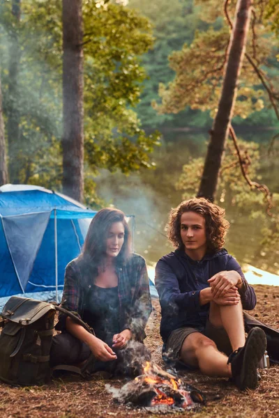 Молодая пара туристов сидит и разговаривает, согреваясь у костра в лагере в лесу. Концепция путешествий, туризма и походов . — стоковое фото