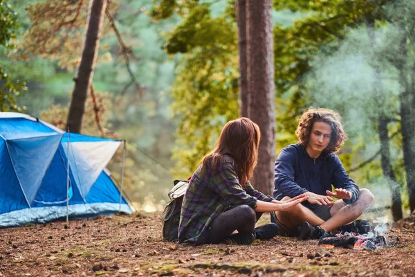 Jovem casal feliz sentado e aquecido perto de uma fogueira no acampamento na floresta . — Fotografia de Stock