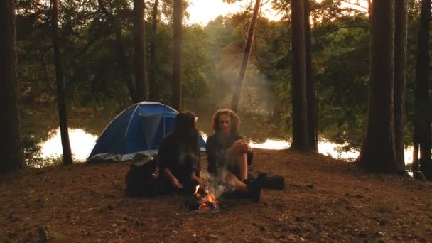 Young couple in the autumn forest in front of fire with camping tent during sunset. — Stock Video