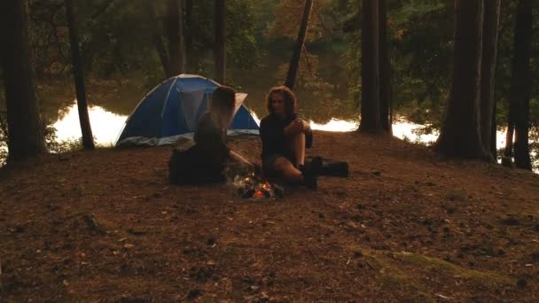 Jeune couple dans la forêt d'automne devant le feu avec tente de camping au coucher du soleil . — Video