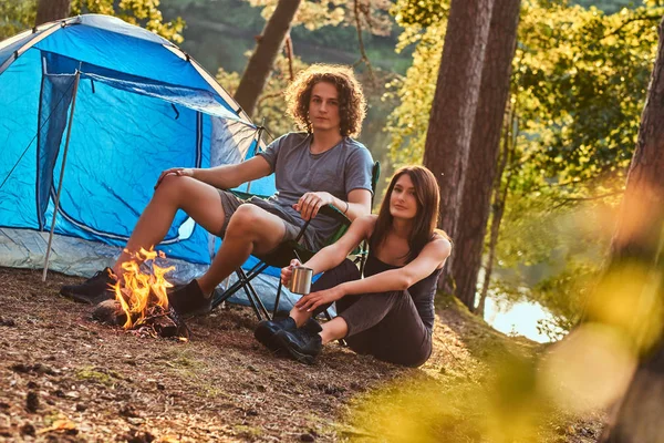 Jovem casal de caminhantes sentados aquecendo perto de uma fogueira no acampamento na floresta no dia ensolarado . — Fotografia de Stock
