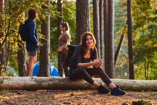 Chica excursionista feliz sentado en el tronco de un árbol y sostiene un teléfono inteligente en el bosque. Viajes, turismo y concepto de caminata . — Foto de Stock