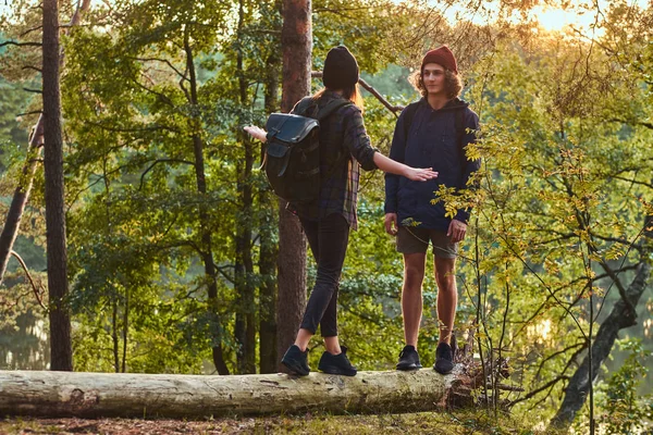 Ein junges Hipster-Paar amüsiert sich bei Sonnenuntergang auf einem Baumstamm in einem wunderschönen Wald. Reise- und Wanderkonzept. — Stockfoto