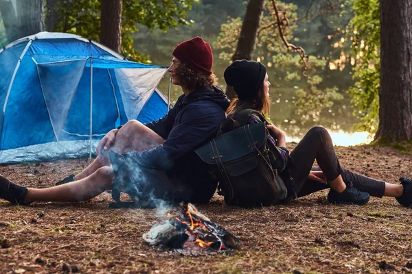 Fiatal csípő pár túrázás az erdőben, és ült vissza, vissza, miközben felmelegedés a következő látnivaló közelében: egy tábortűz a tábor. — Stock Fotó