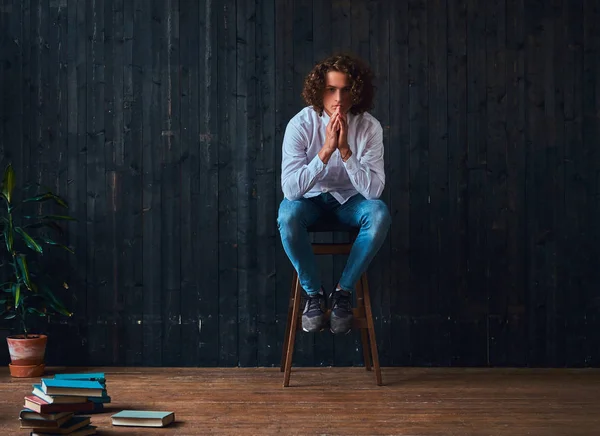 Beau étudiant bouclé tient la main sur le menton tout en étant assis sur une chaise dans une pièce à l'intérieur minimaliste . — Photo