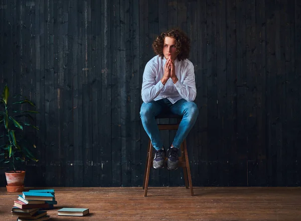 Beau étudiant bouclé tient la main sur le menton tout en étant assis sur une chaise dans une pièce à l'intérieur minimaliste . — Photo