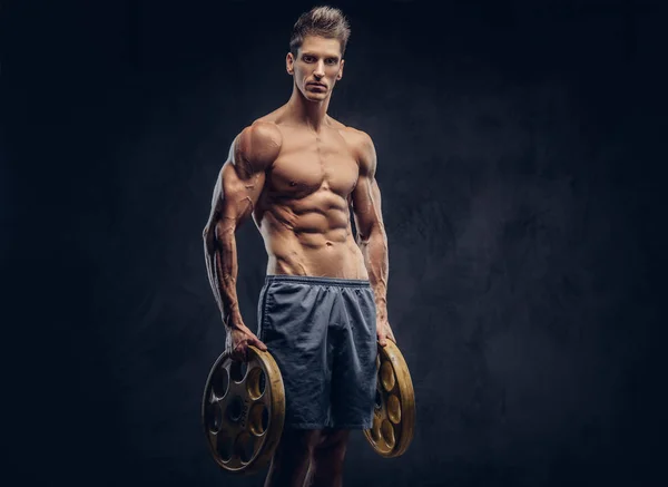 Handsome shirtless ectomorph bodybuilder with stylish hair posing with a barbell disk on dark background. — Stock Photo, Image