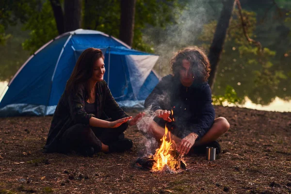 Boldog fiatal pár ül és a felmelegedés egy tábortűz a táborban, az erdő közelében, a naplemente. — Stock Fotó