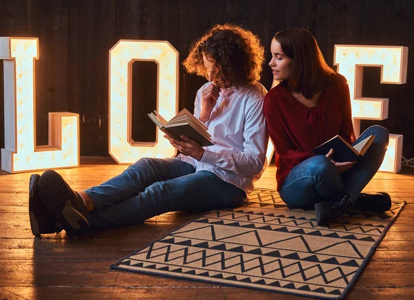 Jeune couple étudiant assis sur un sol et lisant ensemble dans une pièce décorée de lettres volumineuses avec éclairage . — Photo