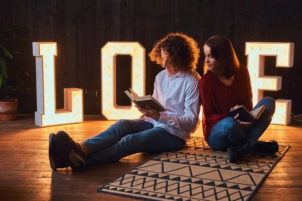 Jeune couple étudiant assis sur un sol et lisant ensemble dans une pièce décorée de lettres volumineuses avec éclairage . — Photo