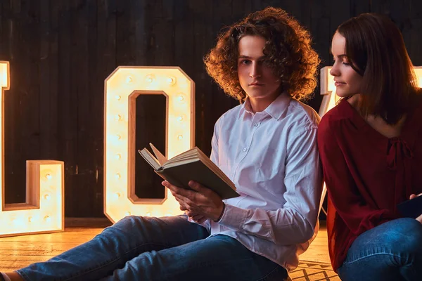 Jovem casal estudante sentado em um piso e lendo juntos em uma sala decorada com letras volumosas com iluminação . — Fotografia de Stock