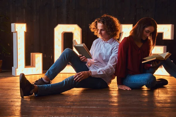 Jeune couple étudiant assis sur un sol et lisant ensemble dans une pièce décorée de lettres volumineuses avec éclairage . — Photo