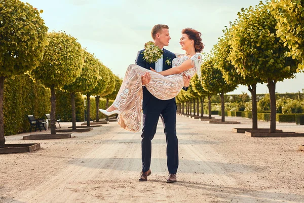 Feliz recém-casados. Bonito noivo mantém sua noiva encantadora em seus braços no jardim incrível . — Fotografia de Stock