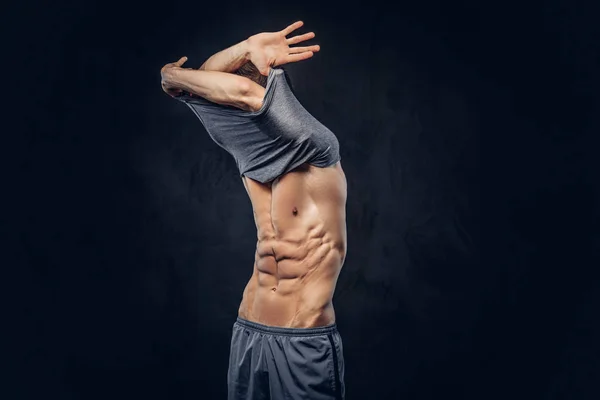 Stylish ectomorph bodybuilder with stylish hair takes off his shirt on the dark background. — Stock Photo, Image