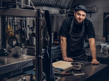 Önlük ve bir masanın üzerine eğilerek ve mutfak kamerada bakarak şapkasını sakallı aşçı.