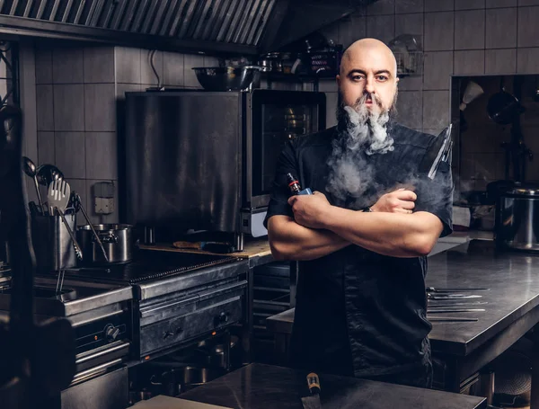 Bearded chef in black uniform smoking e-cigarette while standing in the kitchen. — Stock Photo, Image