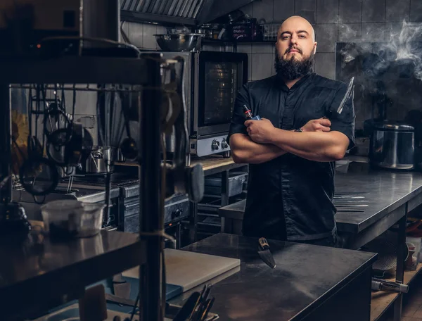 Chef barbudo de uniforme preto fumando e-cigarro enquanto estava na cozinha . — Fotografia de Stock