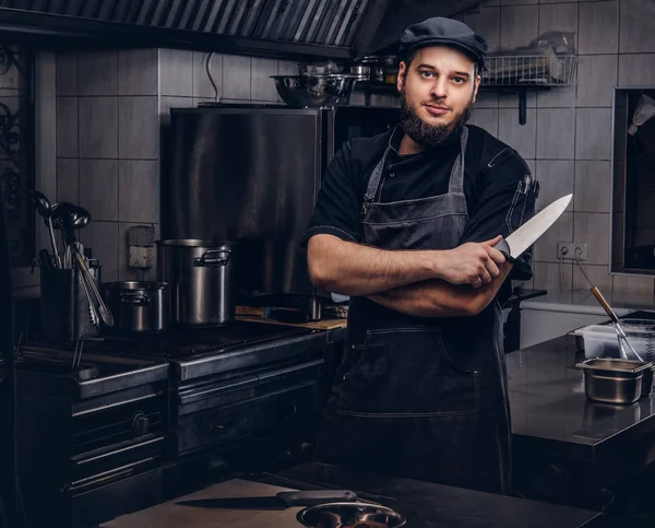 Cozinheiro barbudo em uniforme preto e chapéu segura faca enquanto está de pé com os braços cruzados na cozinha . — Fotografia de Stock