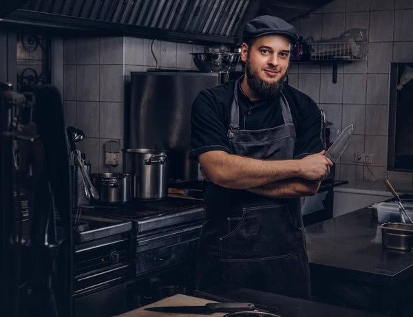 Cocinero barbudo en uniforme negro y sombrero sostiene cuchillo mientras está de pie con los brazos cruzados en la cocina . — Foto de Stock