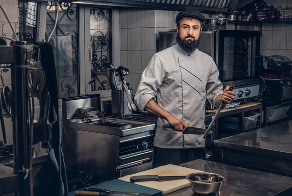 Sakallı aşçı üniforma ve mutfak kamerada bakarak şapka giymiş. — Stok fotoğraf