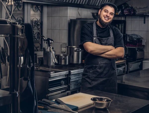 Lächelnder bärtiger Koch in schwarzer Uniform steht mit verschränkten Armen in der Küche. — Stockfoto