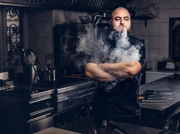 穿着黑色制服的大胡子厨师站在厨房里抽着电子烟. — 图库照片