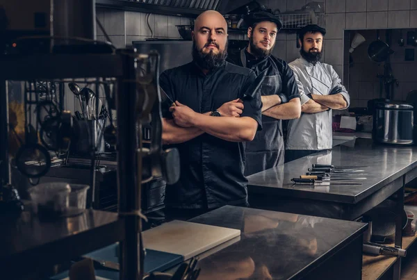 Équipe de cuisiniers barbus professionnels vêtus d'uniformes posant avec des couteaux dans la cuisine . — Photo
