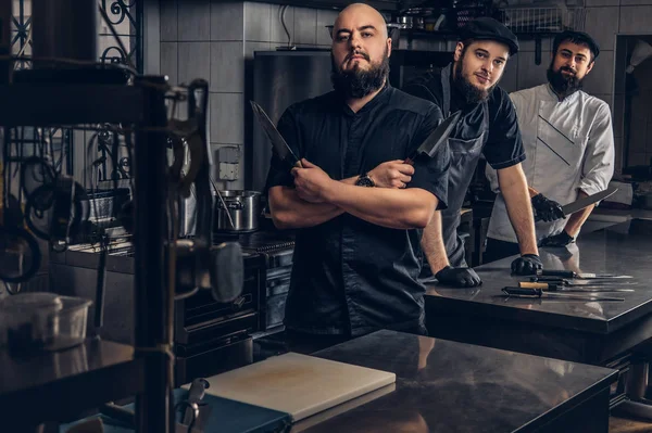 Команда професійних бородатих кухарів, одягнених у форму позування з ножами на кухні . — стокове фото