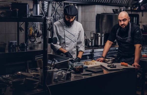 Dos cocineros brutales vestidos con uniformes preparando sushi en una cocina . —  Fotos de Stock