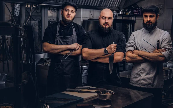 Equipe de cozinheiros barbudos profissionais vestidos com uniformes posando com facas na cozinha . — Fotografia de Stock