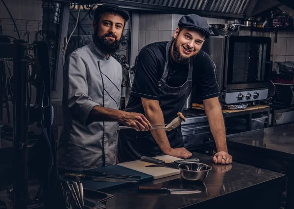 Sorrindo cozinheiros barbudos vestidos com uniformes e chapéus se divertindo na cozinha . — Fotografia de Stock