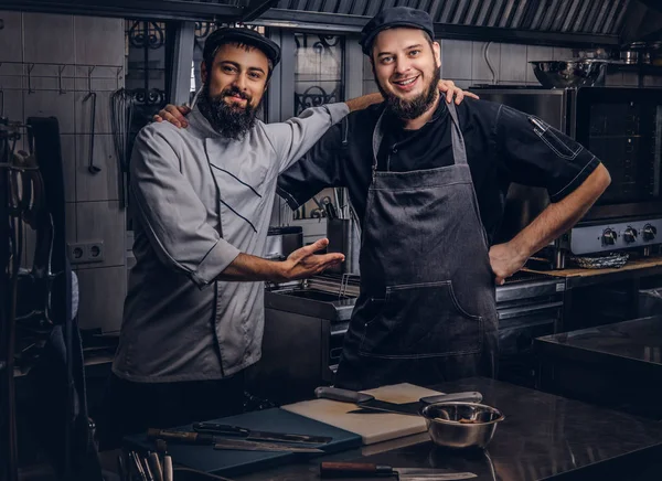 Dwa przyjaciel szczęśliwy Brodaty kucharze ubrani w mundury i czapki objęcia podczas pozowanie w kuchni. — Zdjęcie stockowe