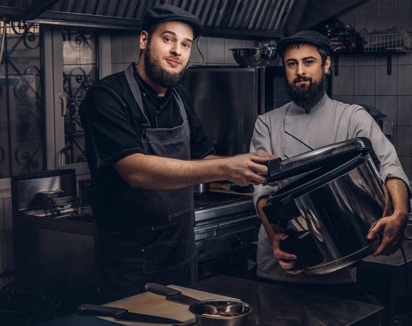 Deux cuisiniers brutaux vêtus d'uniformes et chapeaux debout avec une grande cuisinière dans la cuisine . — Photo
