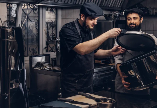 Deux cuisiniers brutaux vêtus d'uniformes et chapeaux debout avec une grande cuisinière dans la cuisine . — Photo