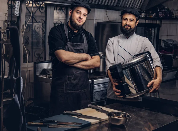 Zwei brutale Köche in Uniformen und Hüten stehen mit großem Multikocher in der Küche. — Stockfoto