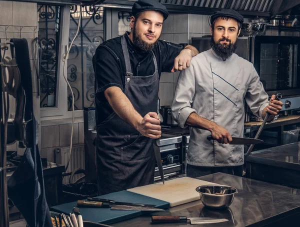 Deux cuisiniers barbus vêtus d'uniformes et chapeaux posant dans la cuisine, regardant une caméra . — Photo